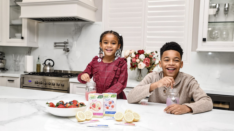 Two children in a white kitchen stirring True Lemon packets into their glasses of water