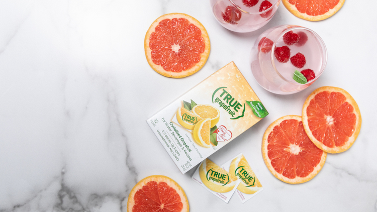 An image of a box of True Grapefruit lying on top of a counter. There are grapefruit slices and two glasses on the counter as well.