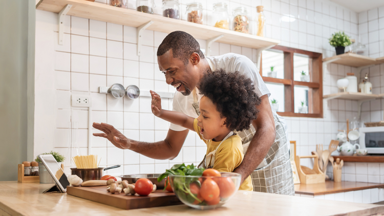 A father an son in the kitchen cooking a meal together. There are vegetables on the counter and they are both laughing. 