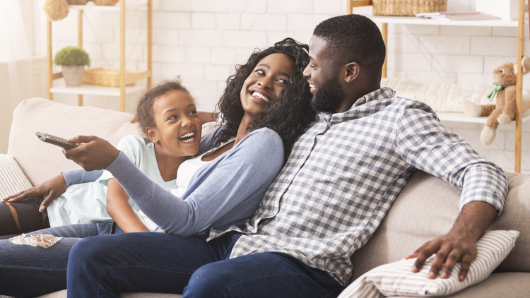 A family of three lounges on the couch together. The father has his arm over the chair's armrest and his wife leans on him with their ten year old kid laying in her lap. They're all laughing. 