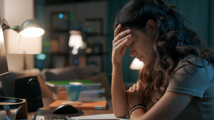 It's night, and a woman is at her desk in her home feeling overwhelmed and exhausted. Her hand is on her forehead and her eyes are closed.