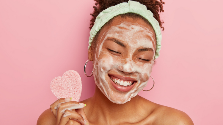 A smiling woman with a green headband has cleanser on her face. She is holding a small pink sponge shaped like a heart. The background is a bright pink. 