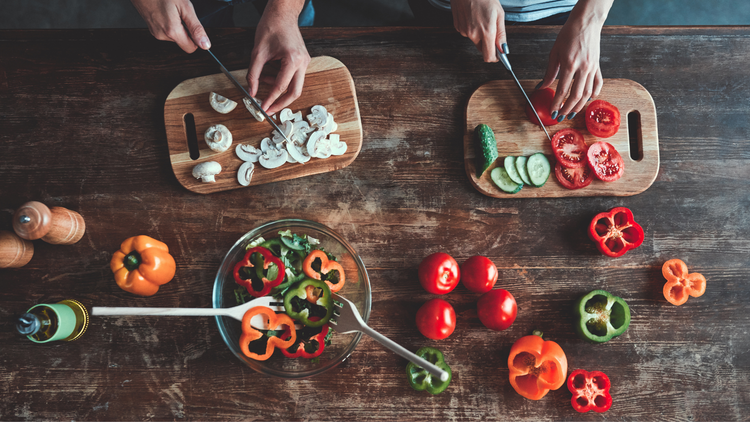 A couple slicing vegetables like bell peppers and onions. The vegetables are bright in color and strewn along the table. 