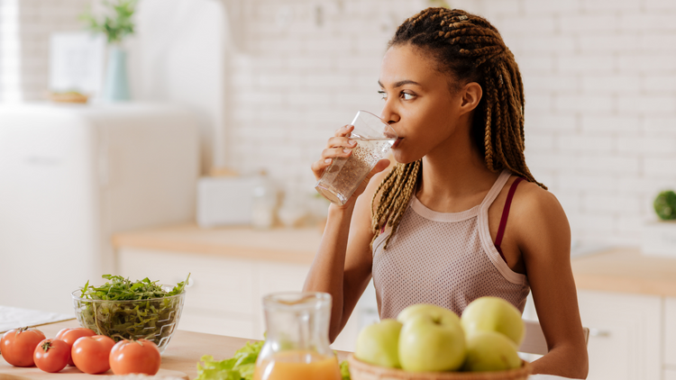 A woman drinking a glass of water in her kitchen as she unpacks groceries.