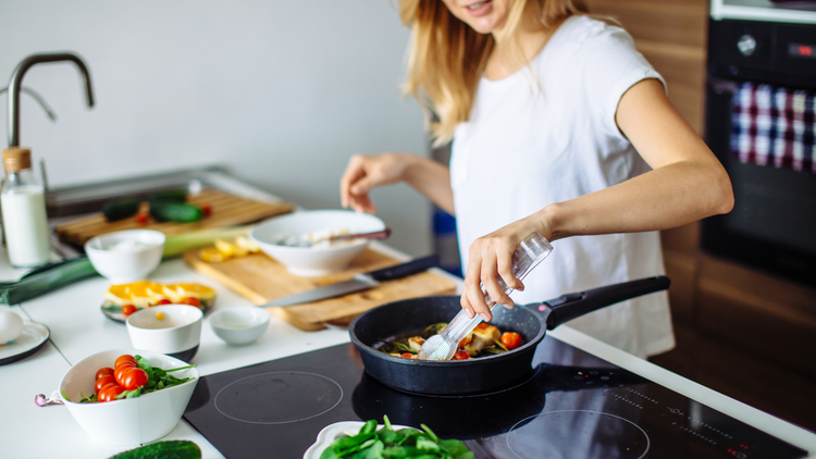 A photo of a woman in a kitchen cooking a meal in a pan, on the counter around her are different ingredients.