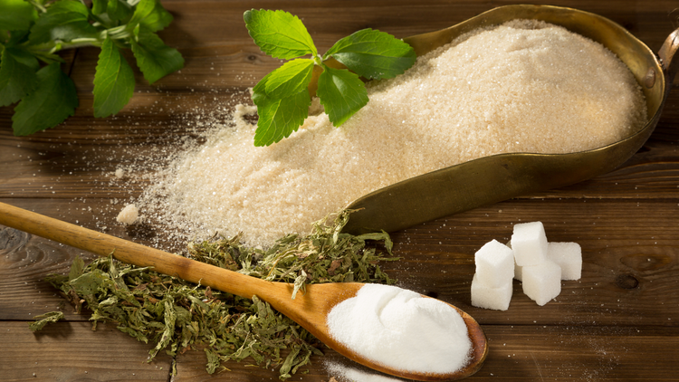 Large scoop filled with Stevia grains, next to a smaller spoon, stevia leaves, and stevia cubes. 