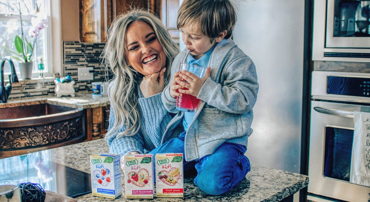 Woman smiles while watching her young son sitting on the kitchen island hugging a glass of True Lemon