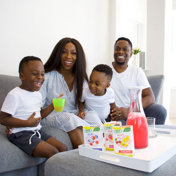 Mother, father, and two boys sitting on grey couch, smiling and sharing a pitcher of True Lemon drink