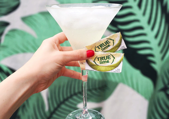 Woman holds cocktail glass and True Lime packets in front of a wall with tropical print wallpaper