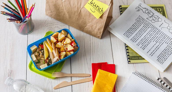 On a white table there is a child's healthy school lunch of sliced fruit with their school books and colored pencils