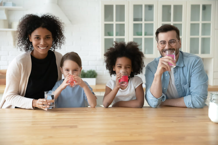 Family in a white kitchen standing behind a wooden kitchen island and facing the camera while drinking glasses of True Lemon drink