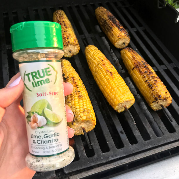 A person holds up a container of True Lime Garlic Cilantro Spice Blend as they grill cobs of corn. 