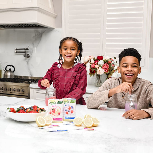 Two kids sit at the kitchen counter and smile while drinking True Lemon Kids. 