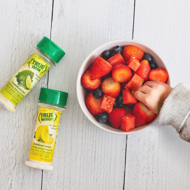 A little kid reaches for a large bowl of fresh strawberries and watermelon, which are lightly dashed with True Lemon. A True Lemon Shaker and True Lime Shaker rest on the table next to the bowl.