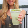 In front of a lake, a woman holds a cup filled with True Lime Original Limeade to the camera. The cup has the text 